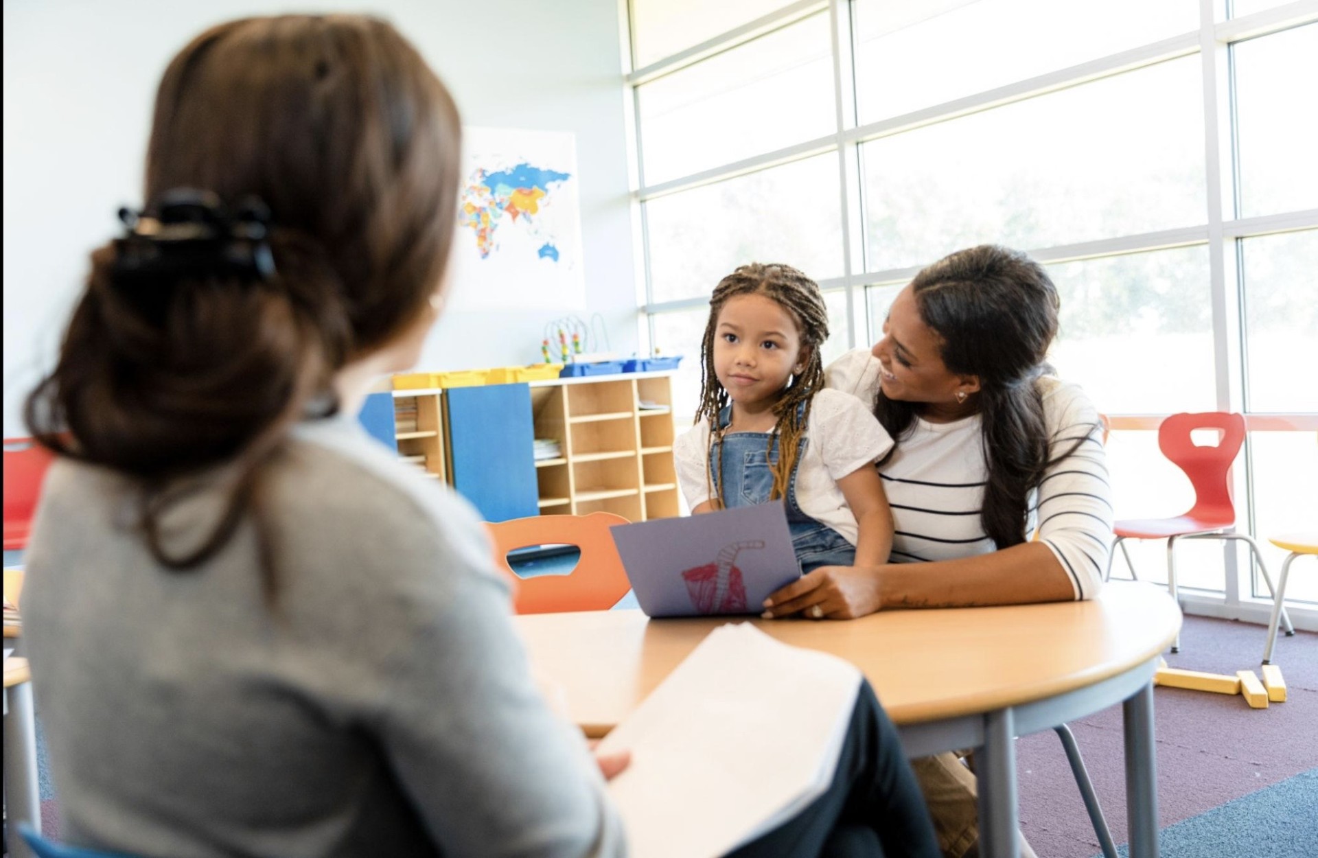 A teacher meets with a parent and child at a table in a bright classroom
